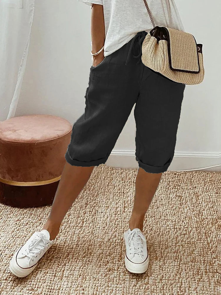 Femme en bermuda noir et t-shirt blanc, portant un sac en osier, debout sur un tapis beige avec des baskets blanches. Mode décontractée chic.