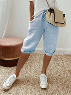 Femme en short bleu clair et t-shirt blanc, portant un sac en osier et des baskets blanches, debout sur un tapis beige. Mode décontractée estivale.