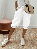 Femme en short blanc décontracté, t-shirt gris, baskets blanches, sac à main beige, posant sur tapis en jute. Mode estivale chic et confortable.