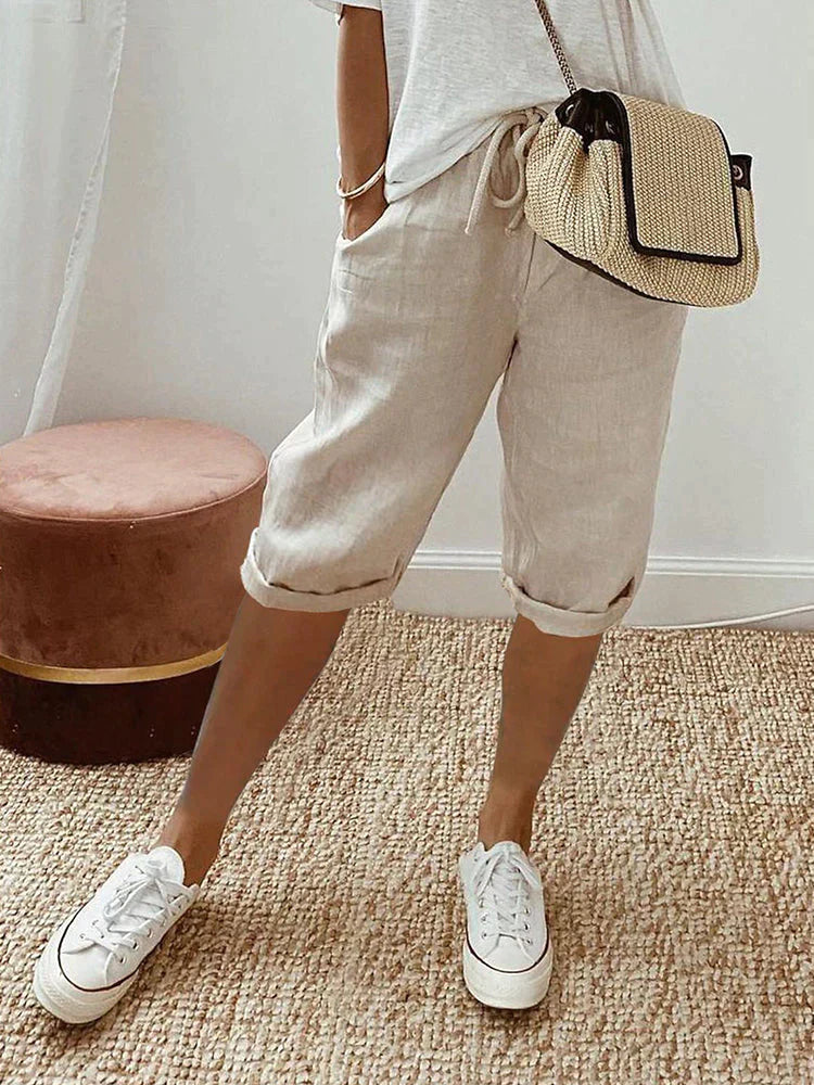 Femme portant un short en lin beige, t-shirt blanc, baskets blanches et sac à main en paille, posant sur un tapis en jute. Mode décontractée estivale.