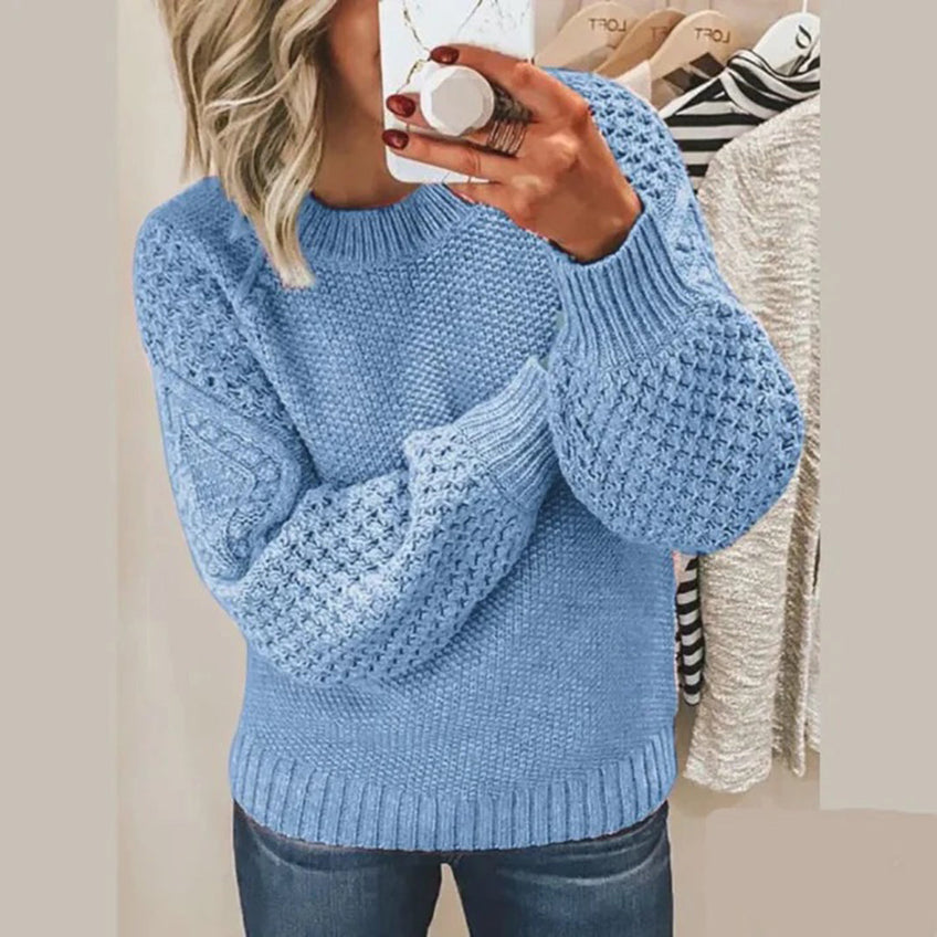 Femme portant un pull bleu en tricot torsadé, manches bouffantes, tenant un smartphone devant un miroir. Mode automne-hiver, style décontracté.