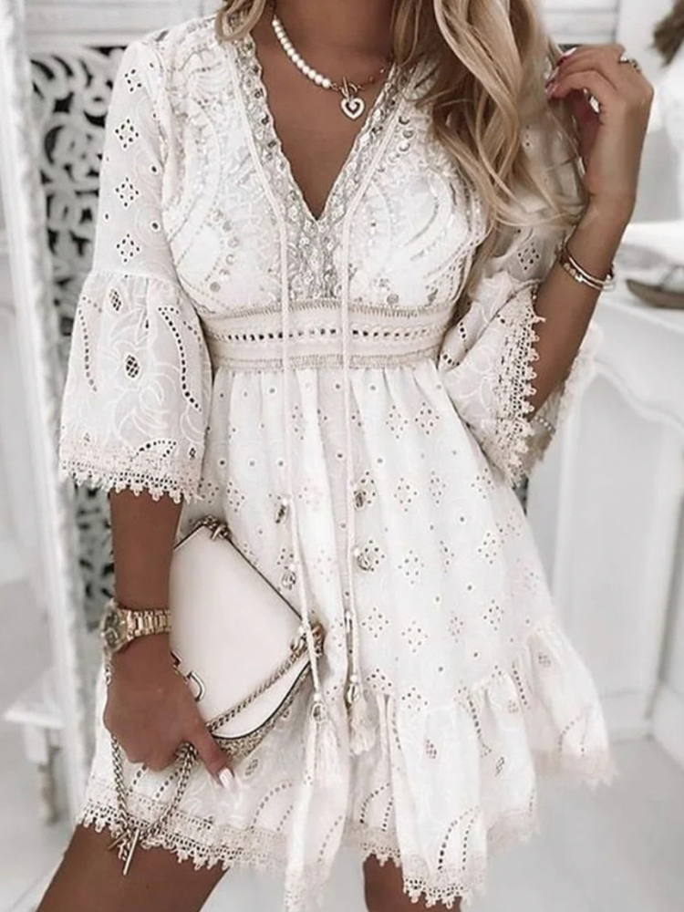 Robe blanche bohème chic en dentelle avec manches évasées, détails brodés, portée par une femme blonde, accessoirisée d'un sac à main et bijoux.