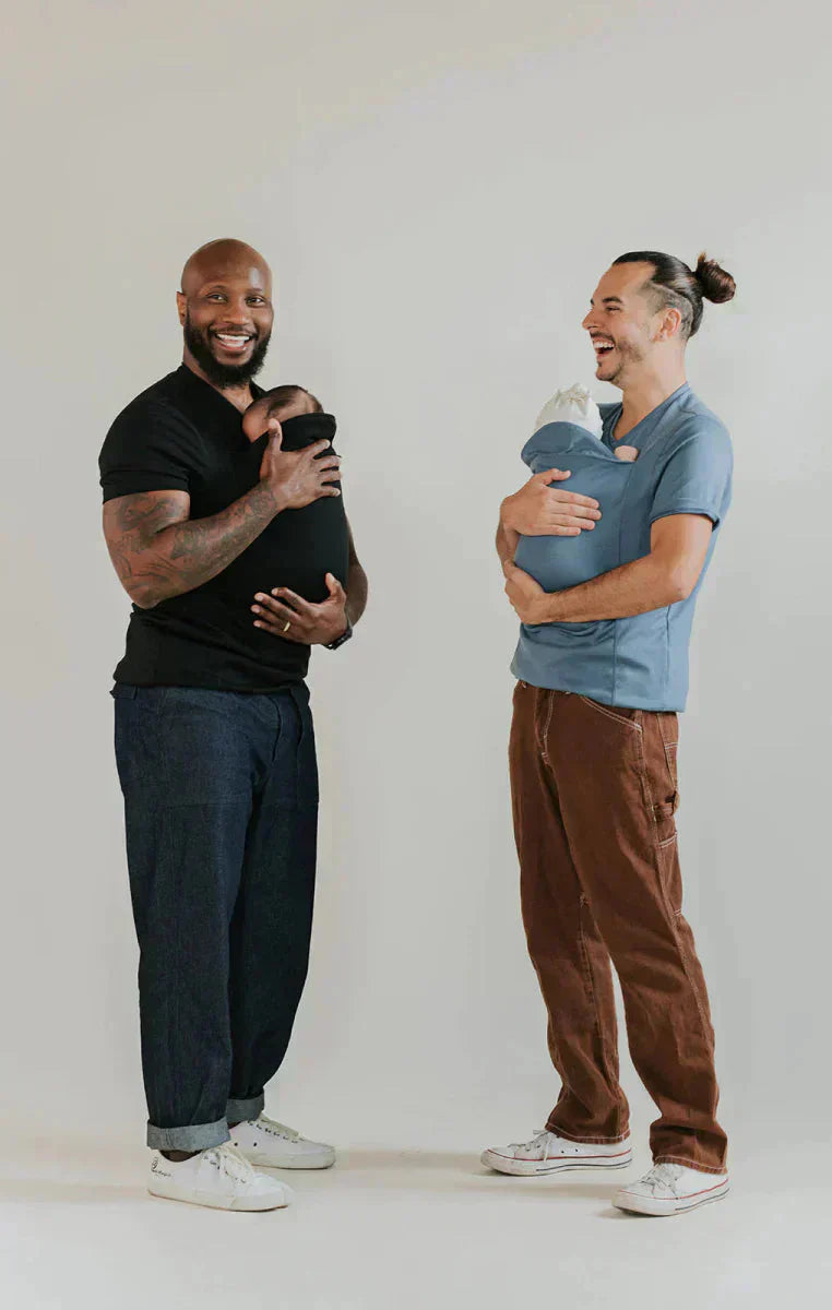 Deux hommes souriants tenant des bébés emmaillotés, l'un en t-shirt noir et l'autre en bleu, sur fond neutre. Paternité, diversité, bonheur.