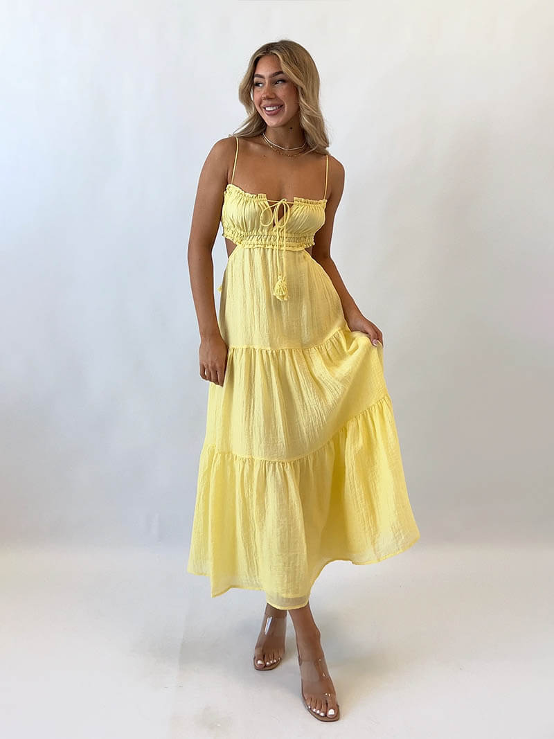 Femme souriante portant une robe longue jaune à volants, bretelles fines, posant sur fond blanc. Mode estivale, tenue élégante et décontractée.