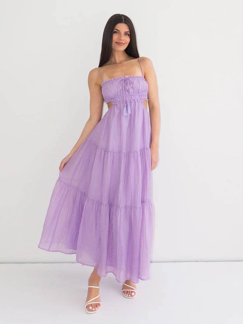 Robe longue violette élégante portée par une femme souriante, avec des bretelles fines et des sandales blanches, sur fond blanc minimaliste.
