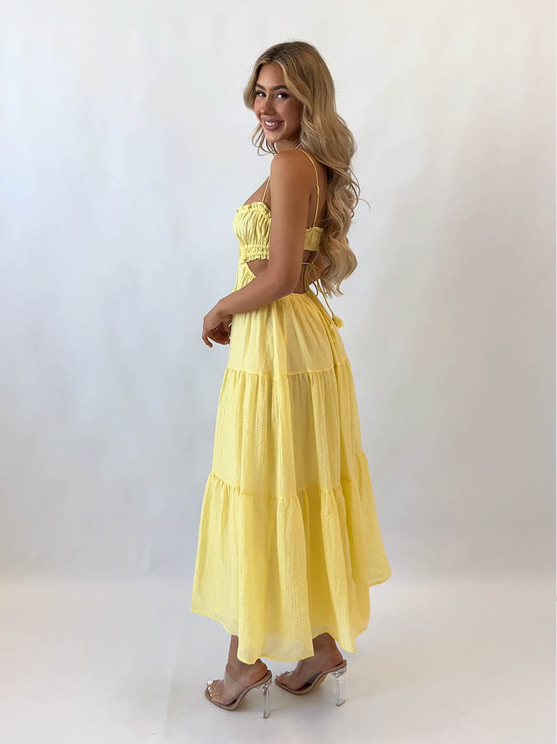 Femme souriante en robe longue jaune, dos nu, posant de profil sur fond blanc. Mode estivale, élégance, style décontracté.