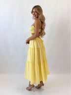 Femme souriante en robe longue jaune, dos nu, posant de profil sur fond blanc. Mode estivale, élégance, style décontracté.