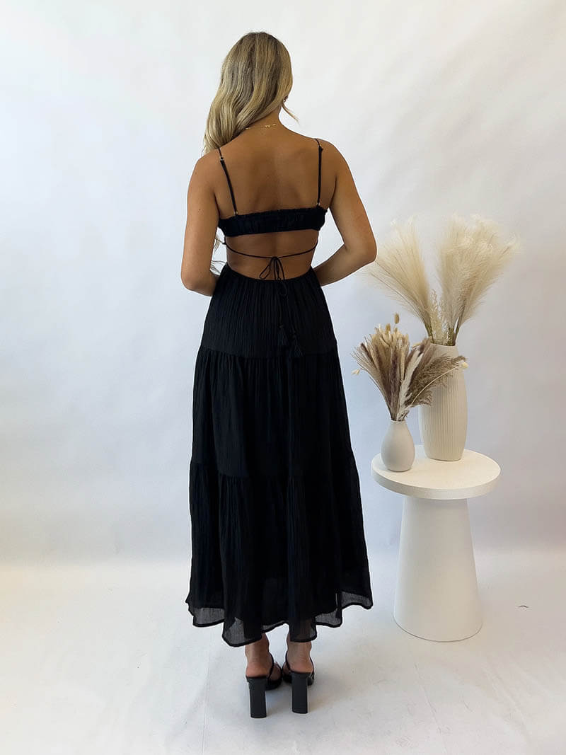 Robe longue noire dos nu, femme blonde de dos, talons hauts, décor minimaliste, vases avec herbes séchées, mode élégante et moderne.