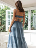 Femme en robe longue bleue dos nu, debout dans une véranda lumineuse avec vue sur jardin. Mode estivale, élégance décontractée, style bohème.