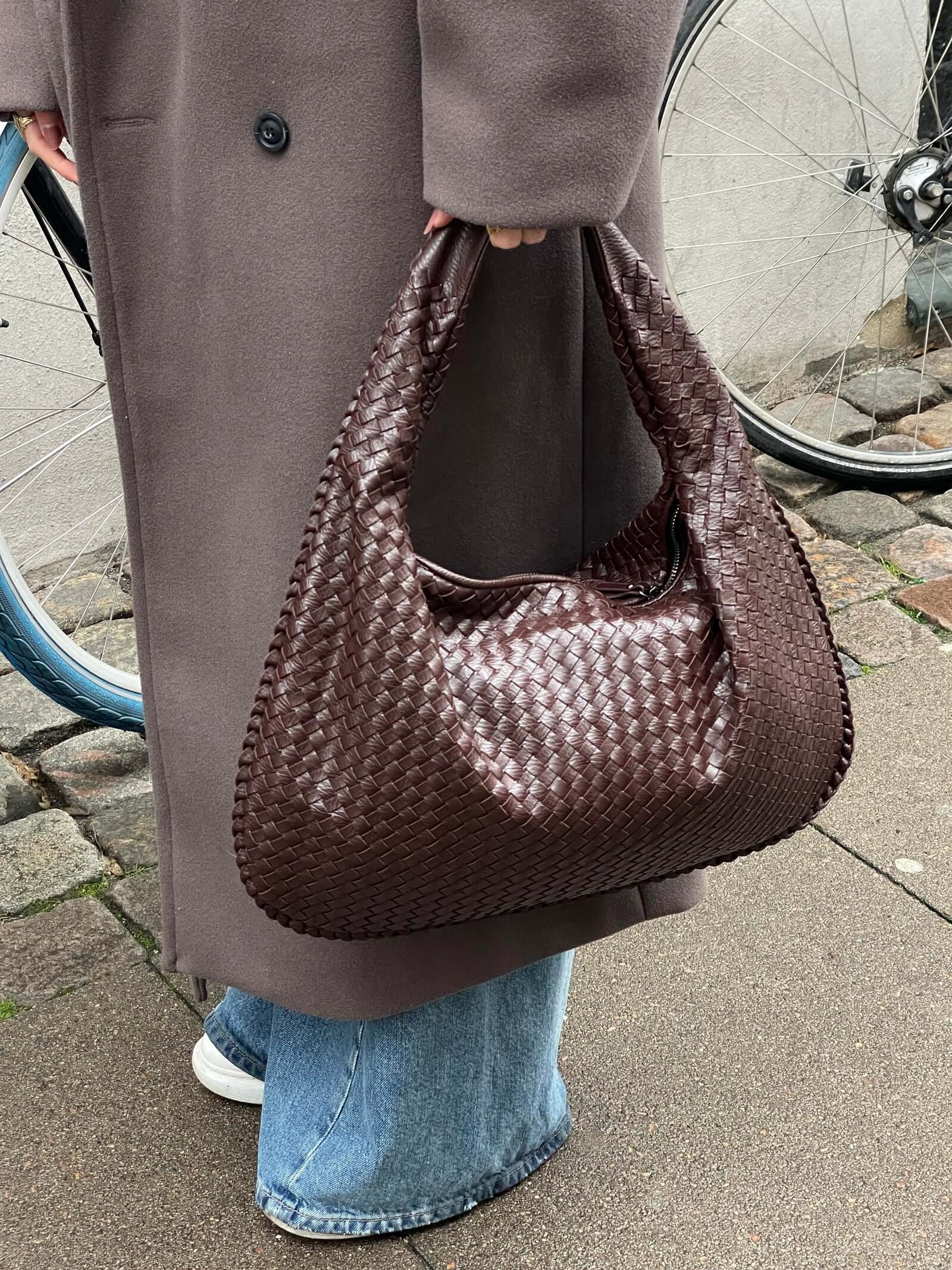 Sac à main en cuir tressé marron porté par une personne en manteau gris et jean, avec une roue de vélo en arrière-plan.