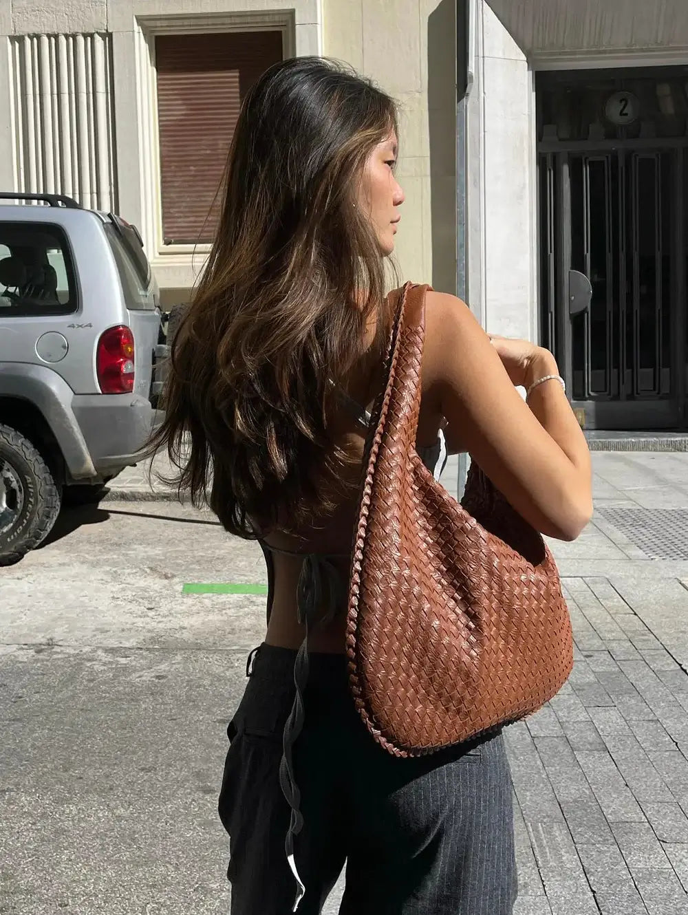 Femme de dos avec un grand sac à main en cuir tressé marron, marchant en ville. Mode urbaine, accessoires tendance, style décontracté.