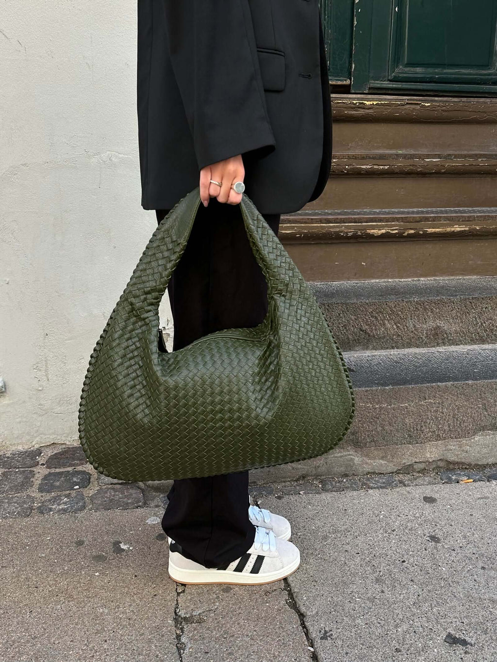Sac à main vert tressé porté par une personne en tenue noire, debout sur un trottoir. Chaussures blanches et noires visibles. Mode urbaine élégante.