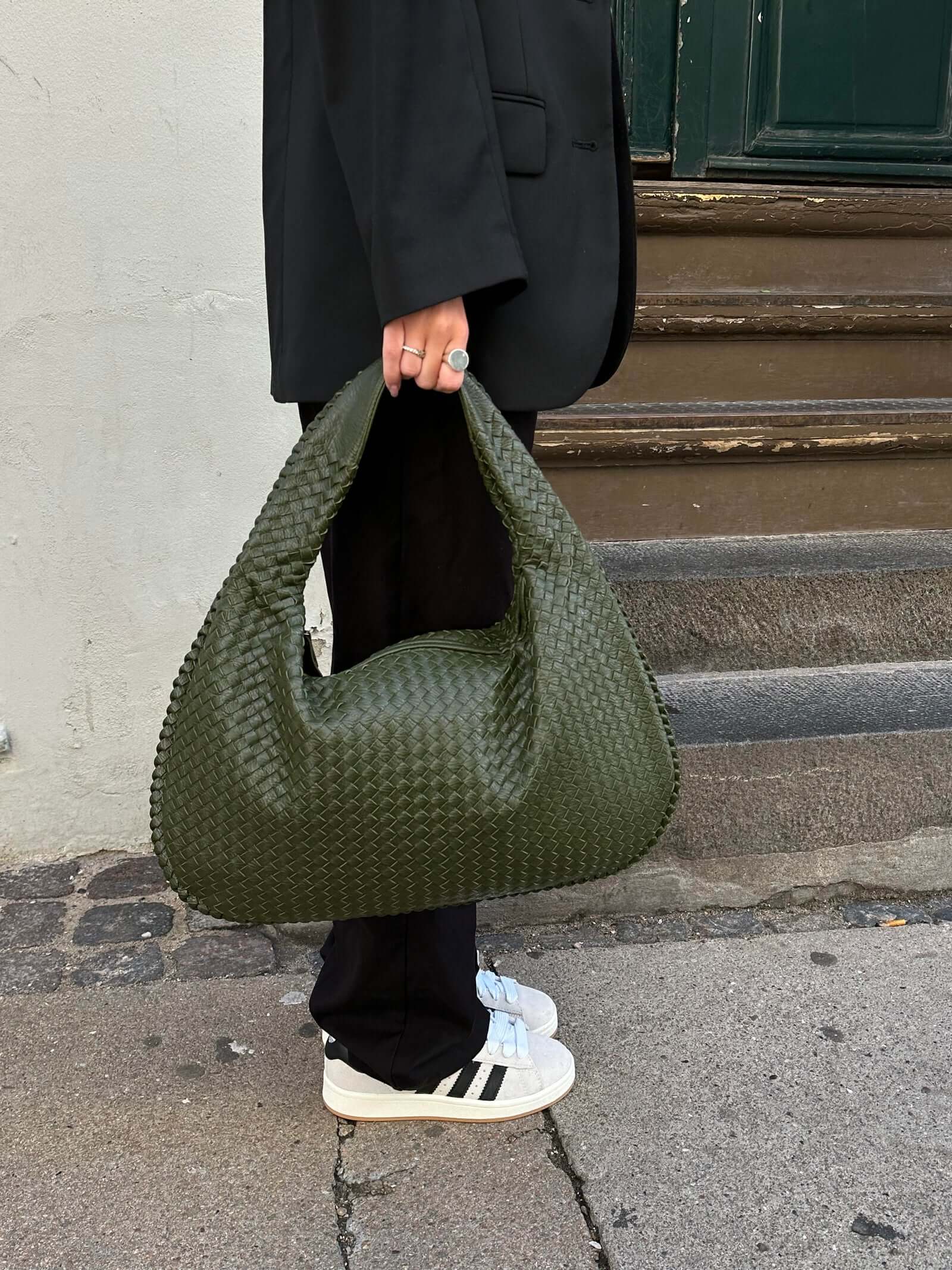 Sac à main vert tressé porté par une personne en tenue noire, debout sur un trottoir. Chaussures blanches et noires visibles. Mode urbaine élégante.