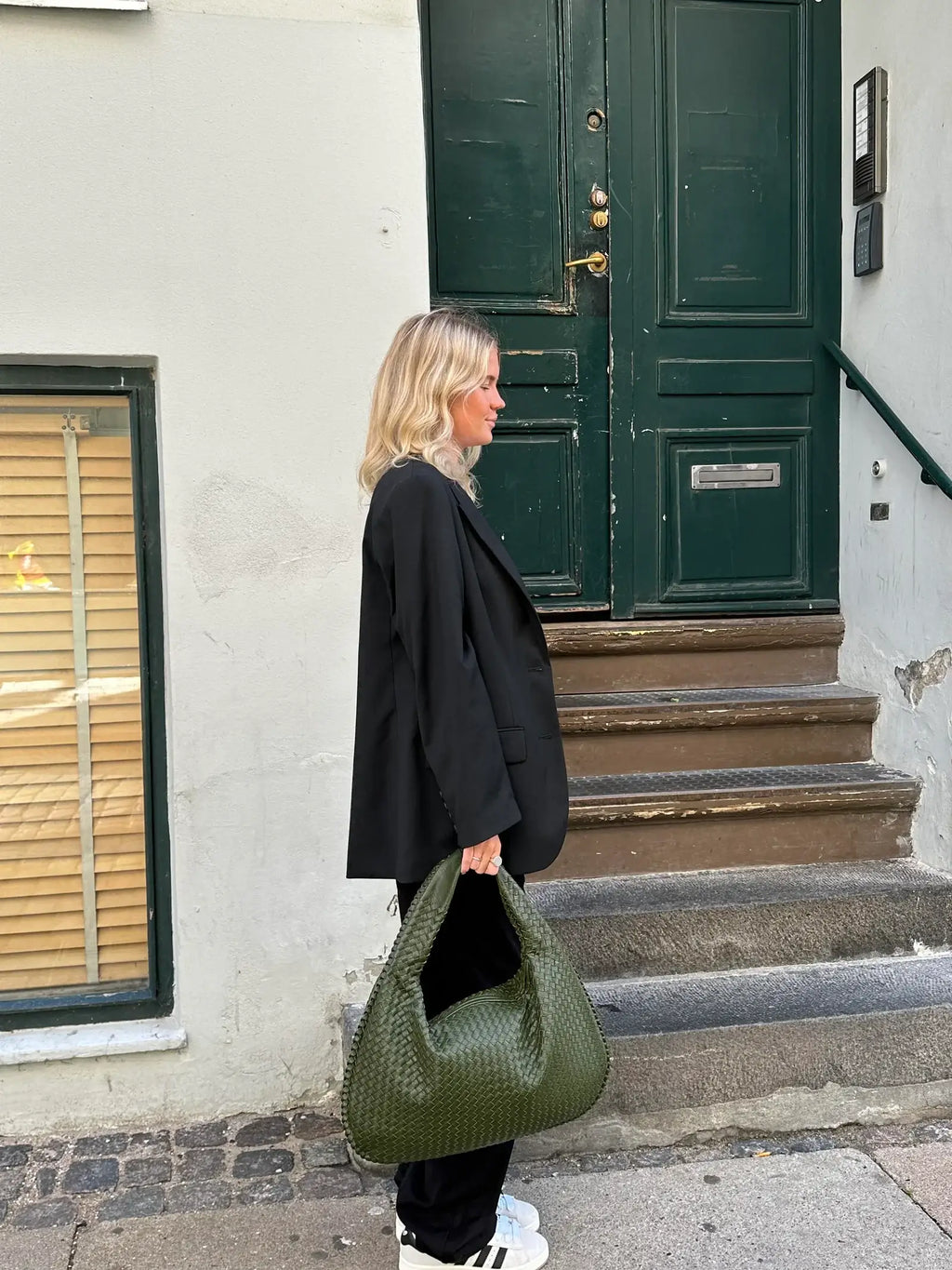 Femme en blazer noir tenant un grand sac vert, debout devant une porte verte. Mode urbaine, style décontracté, tendance automne.