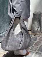 Sac à main tressé gris porté par une personne en manteau gris et pantalon blanc, sur fond de rue pavée. Mode urbaine, accessoire tendance.