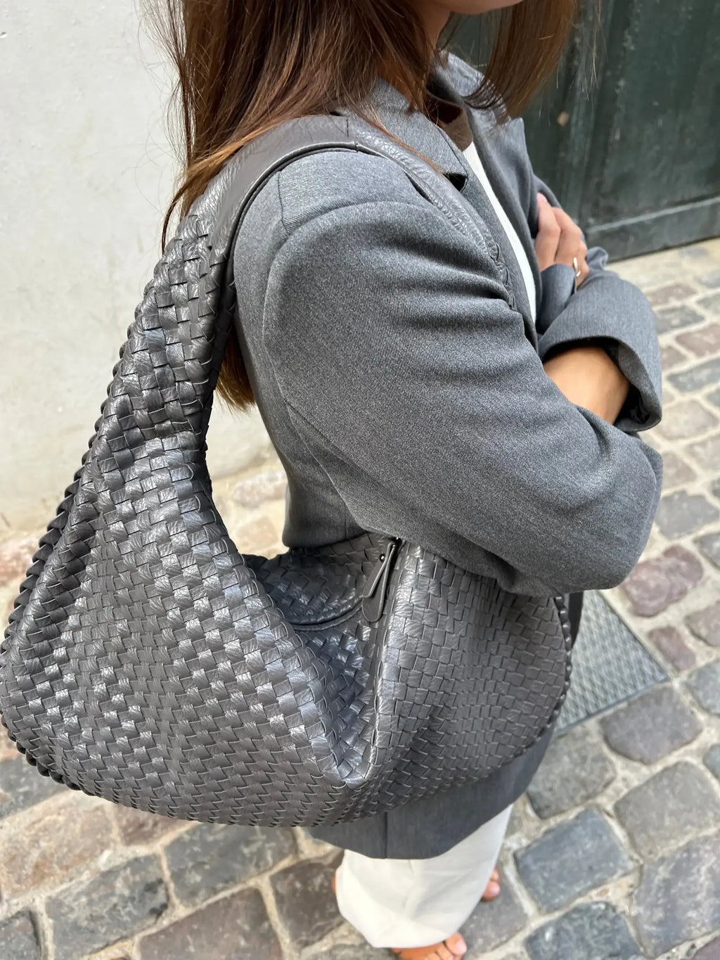Femme portant un sac à main tressé gris, veste grise, sur rue pavée. Mode élégante, accessoires tendance, style urbain chic.