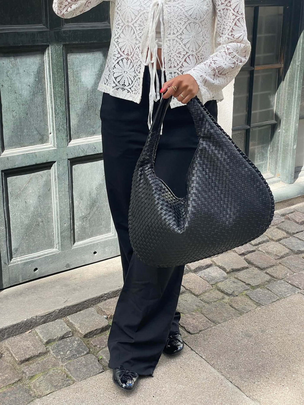 Femme portant un sac à main noir tressé, vêtue d'un chemisier en dentelle blanche et d'un pantalon noir, posant devant une porte en bois.