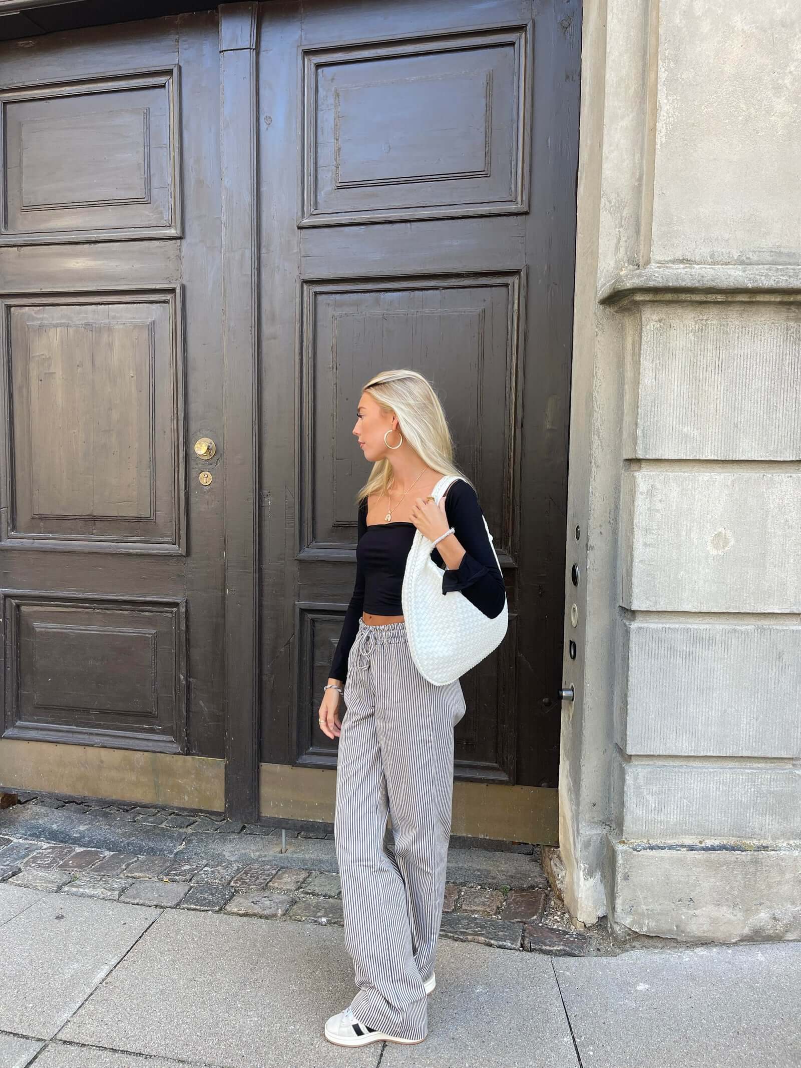 Jeune femme blonde en tenue décontractée, portant un sac blanc, devant une grande porte en bois sombre. Mode urbaine, style moderne, rue pavée.