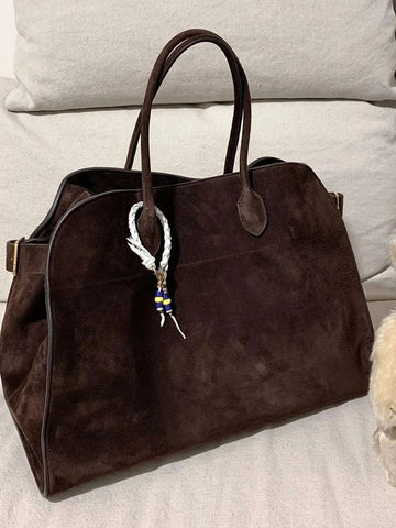 Sac à main en daim marron foncé avec poignées, orné d'un pendentif tressé blanc et bleu, posé sur un canapé beige. Accessoire mode élégant.