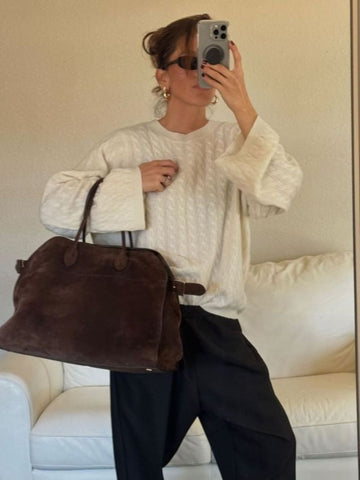 Femme en pull blanc texturé, pantalon noir, portant un grand sac marron en daim, prenant un selfie avec un smartphone devant un canapé blanc.