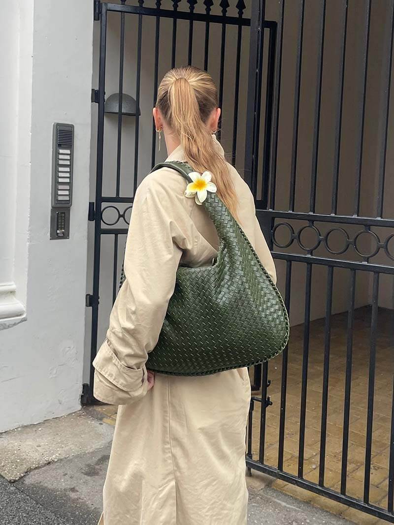 Femme portant un manteau beige et un sac vert tressé avec une fleur blanche, devant une grille noire. Mode urbaine élégante et tendance.