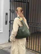 Femme portant un manteau beige et un sac vert tressé avec une fleur blanche, devant une grille noire. Mode urbaine élégante et tendance.