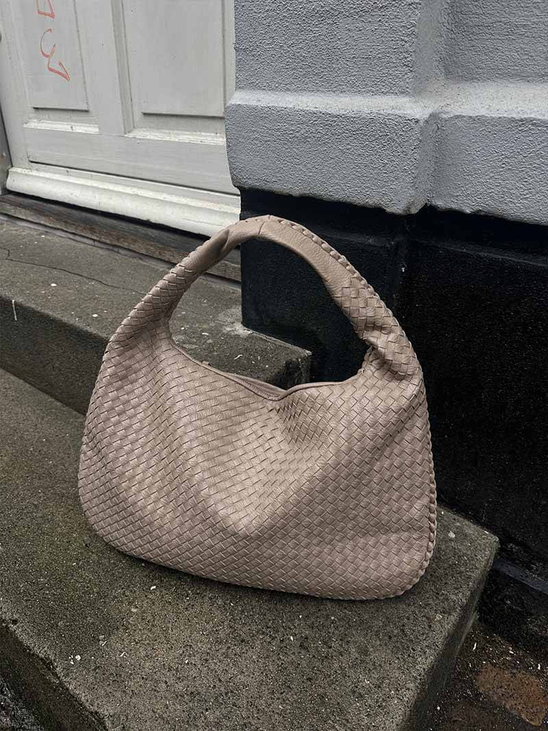 Sac à main en cuir tressé beige posé sur un trottoir en béton devant une porte blanche. Accessoire de mode élégant et tendance.