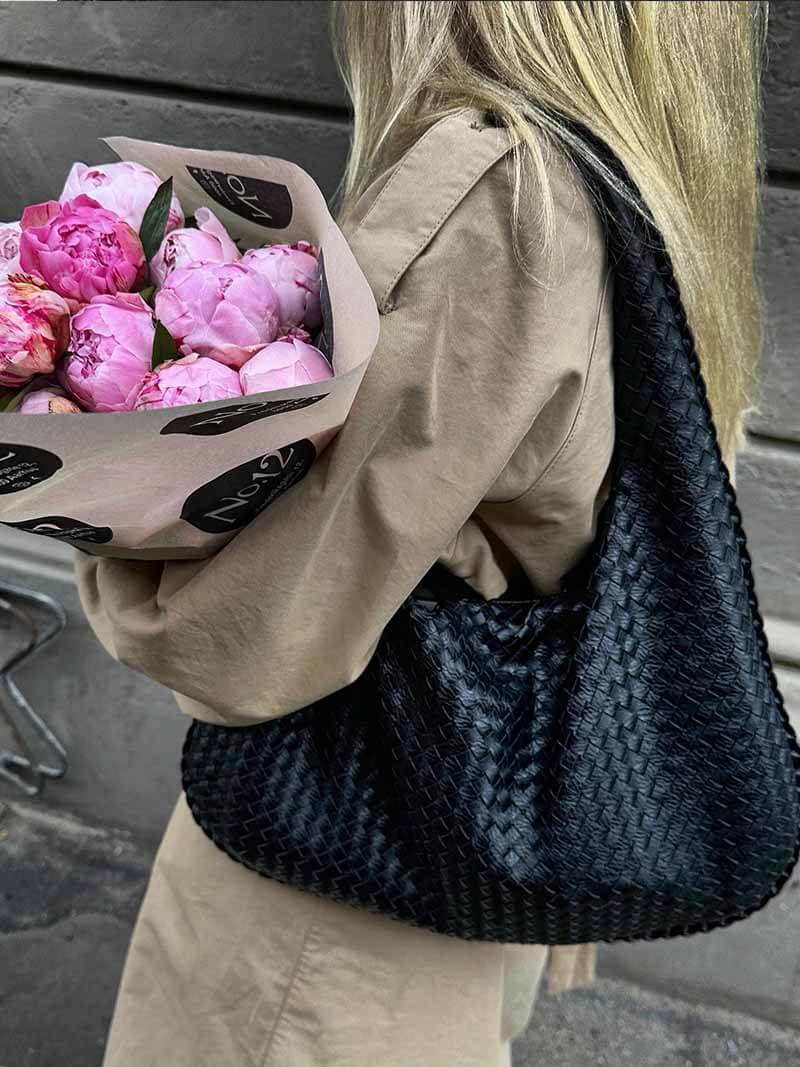 Femme en manteau beige tenant un bouquet de pivoines roses et un sac noir tressé. Mode élégante, accessoires tendance, fleurs fraîches.