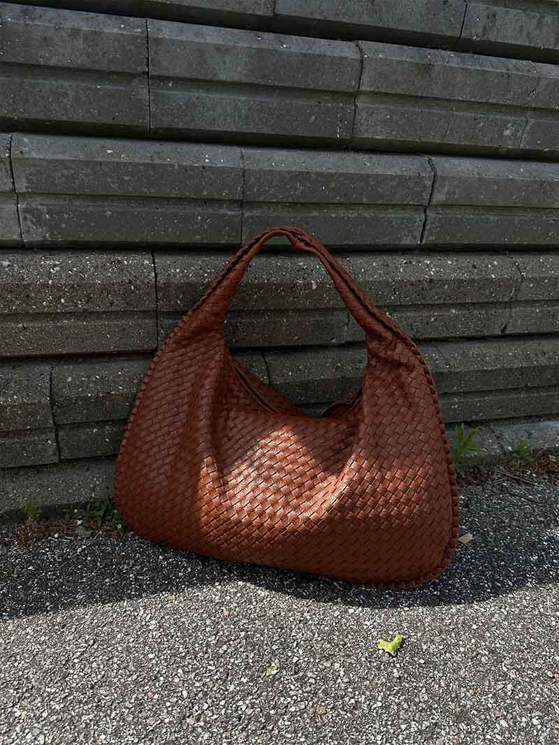Sac à main en cuir tressé marron posé sur un sol en béton devant un mur de briques grises. Accessoire mode élégant et tendance.