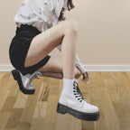 Femme portant des bottes blanches à lacets, jupe noire et chemise blanche, posant sur un sol en bois. Mode tendance, style urbain, chaussures élégantes.