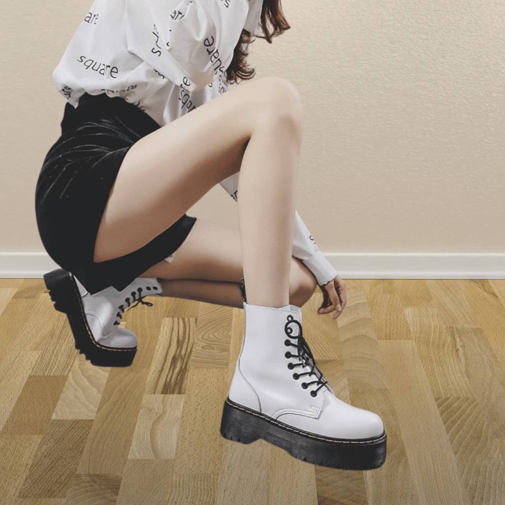 Femme portant des bottes blanches à lacets, jupe noire et chemise blanche, posant sur un sol en bois. Mode tendance, style urbain, chaussures élégantes.