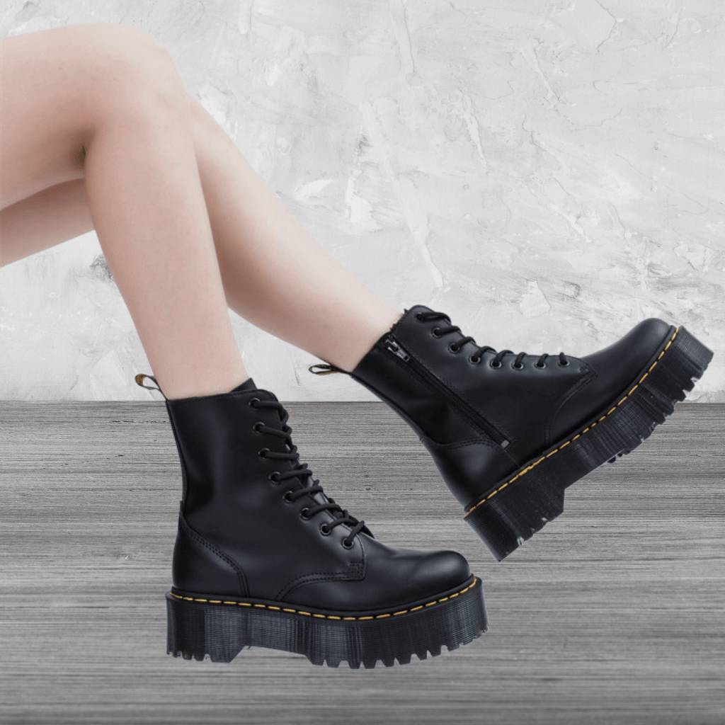 Bottes noires en cuir à semelles épaisses, lacets et fermeture éclair, portées par une personne sur fond de mur texturé gris.