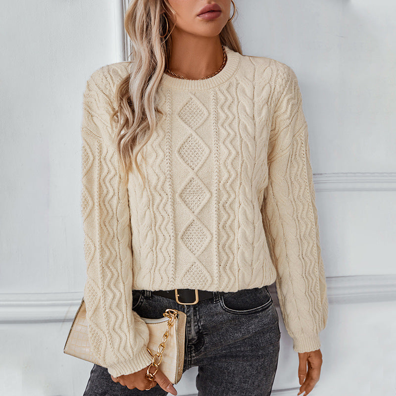 Femme portant un pull en maille torsadée beige, tenant un sac à main beige, posant contre un mur blanc. Mode automne, style décontracté chic.
