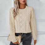 Femme portant un pull en maille torsadée beige, tenant un sac à main beige, posant contre un mur blanc. Mode automne, style décontracté chic.