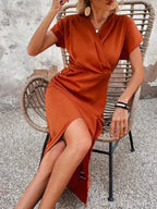 Robe orange élégante, manches courtes, fendue, portée par une femme assise sur une chaise en rotin. Mode féminine chic, tendance été.