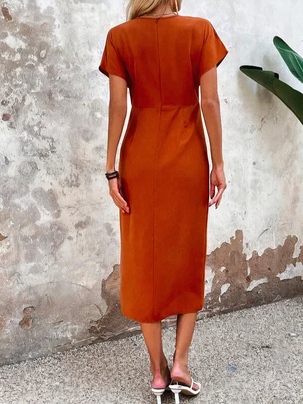 Robe orange élégante vue de dos, femme debout contre un mur texturé, plante verte à droite, style décontracté chic, mode féminine tendance.