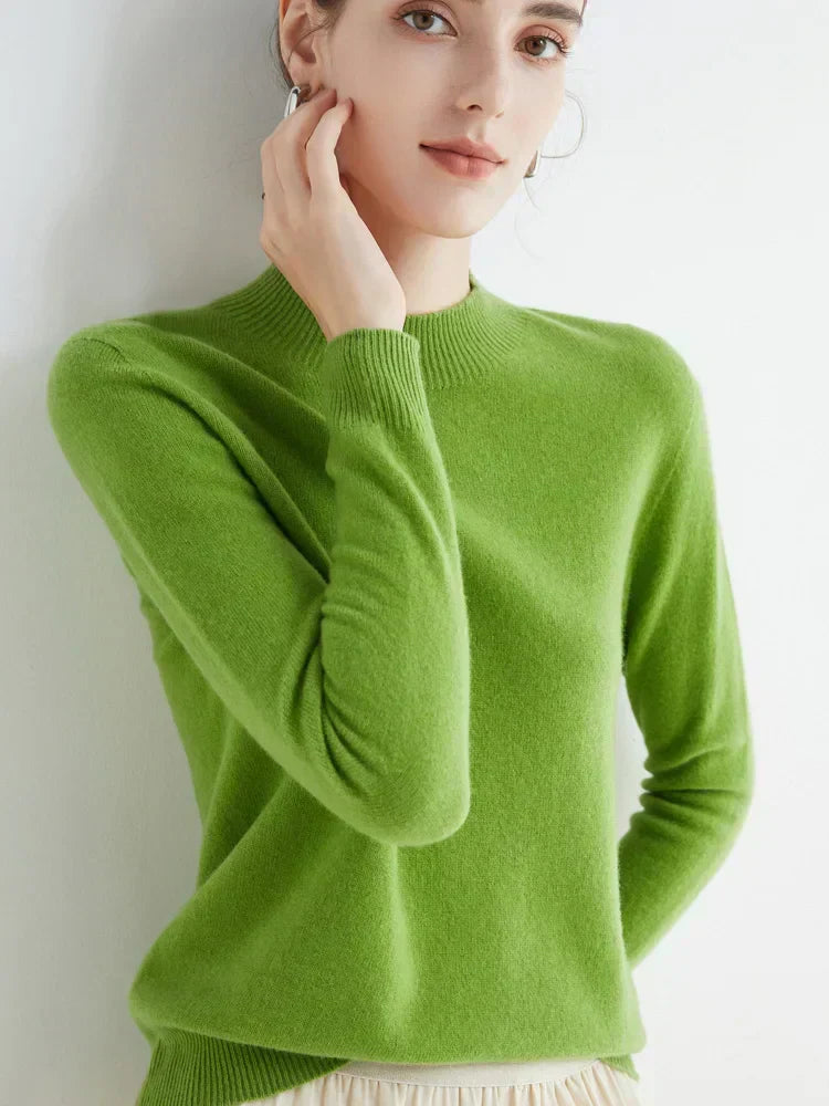 Femme portant un pull vert en cachemire, manches longues, col rond, posant contre un mur blanc. Mode élégante, tenue automne-hiver.