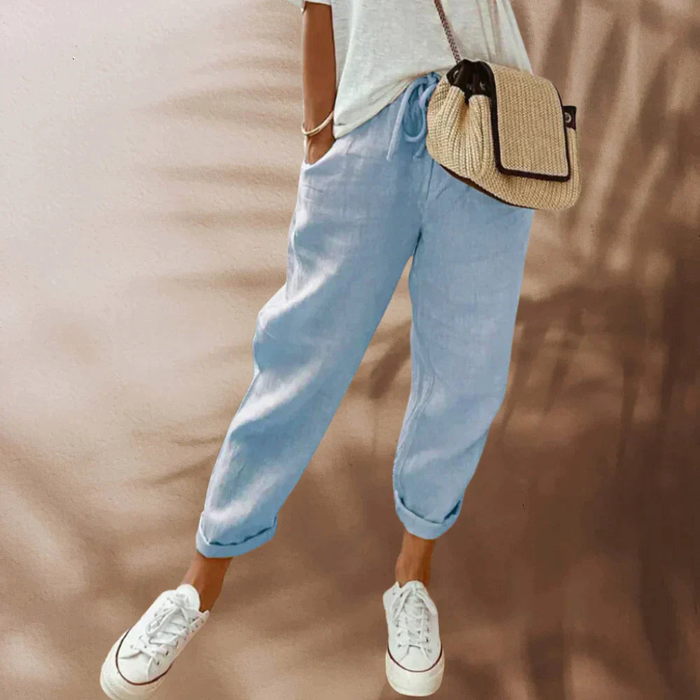 Pantalon en lin bleu clair, décontracté, avec cordon de serrage, porté avec un t-shirt blanc, sac en osier et baskets blanches. Mode estivale tendance.