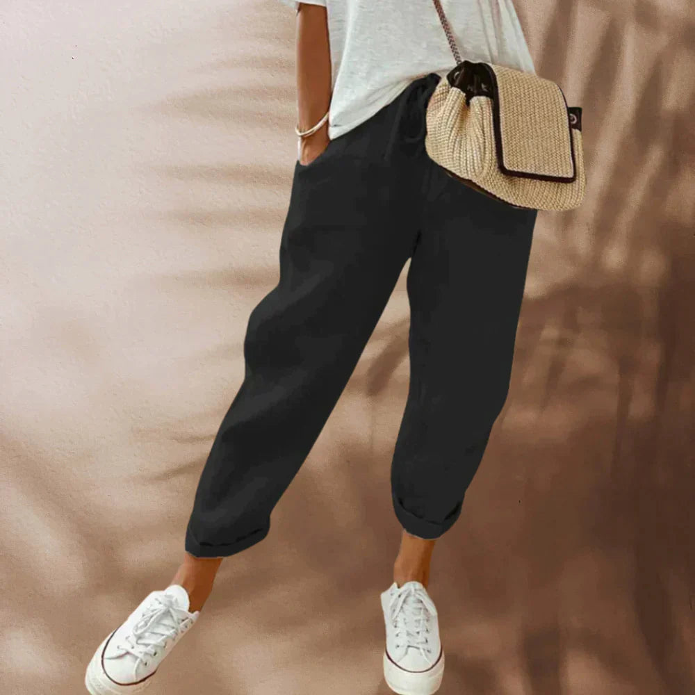 Femme portant un pantalon noir décontracté, t-shirt blanc, baskets blanches et sac en osier. Mode féminine, tenue décontractée, style estival.
