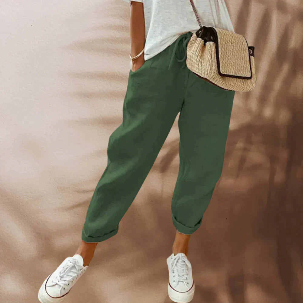 Femme portant un pantalon vert ample, t-shirt blanc, baskets blanches et sac à bandoulière en paille. Mode décontractée, tendance estivale.