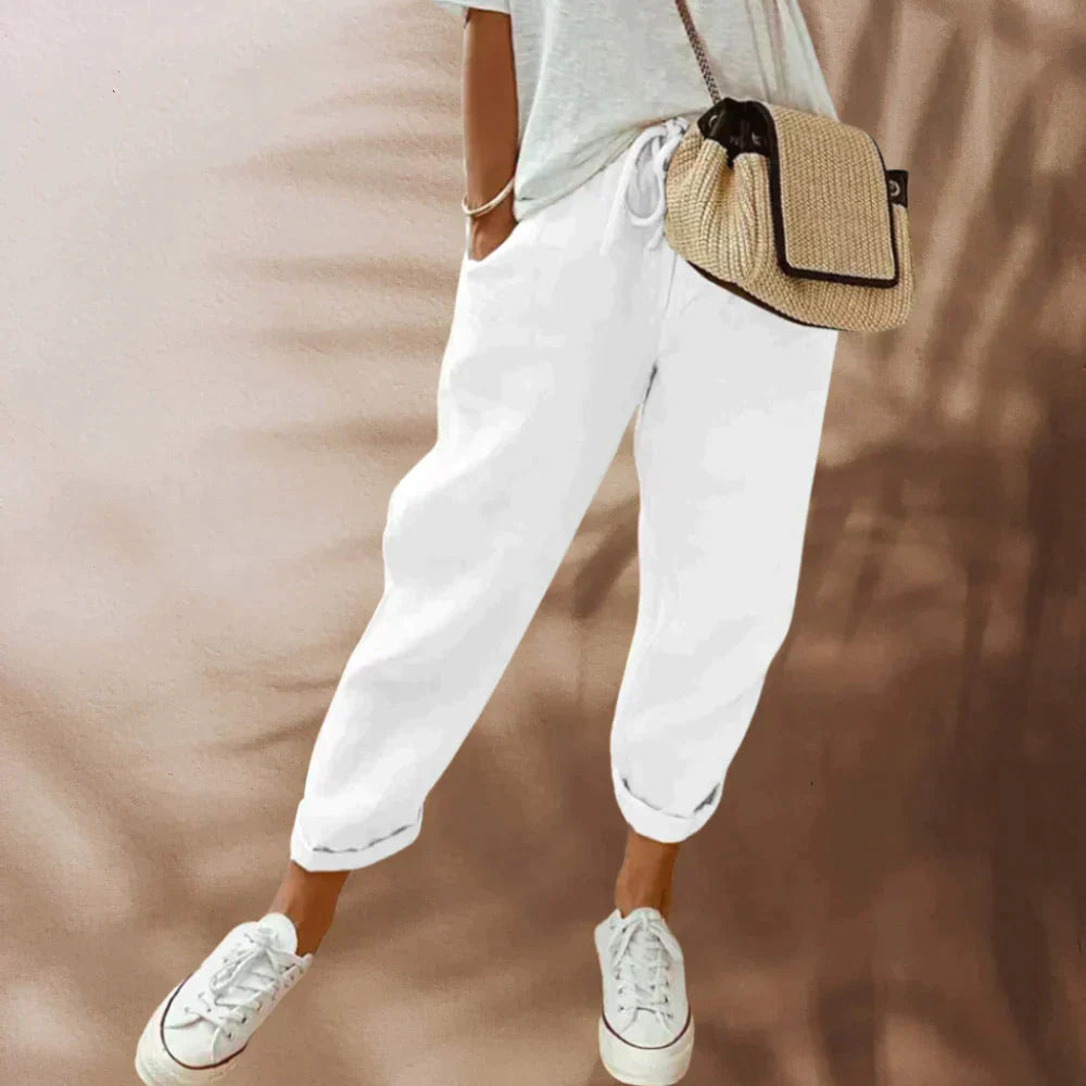 Femme en pantalon blanc décontracté, t-shirt gris, baskets blanches et sac en paille. Mode estivale, tenue confortable et tendance.