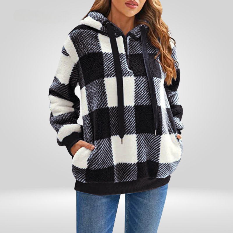 Femme portant un sweat à capuche à carreaux noir et blanc, manches longues, poches avant, style décontracté, sur fond gris. Mode automne-hiver tendance.