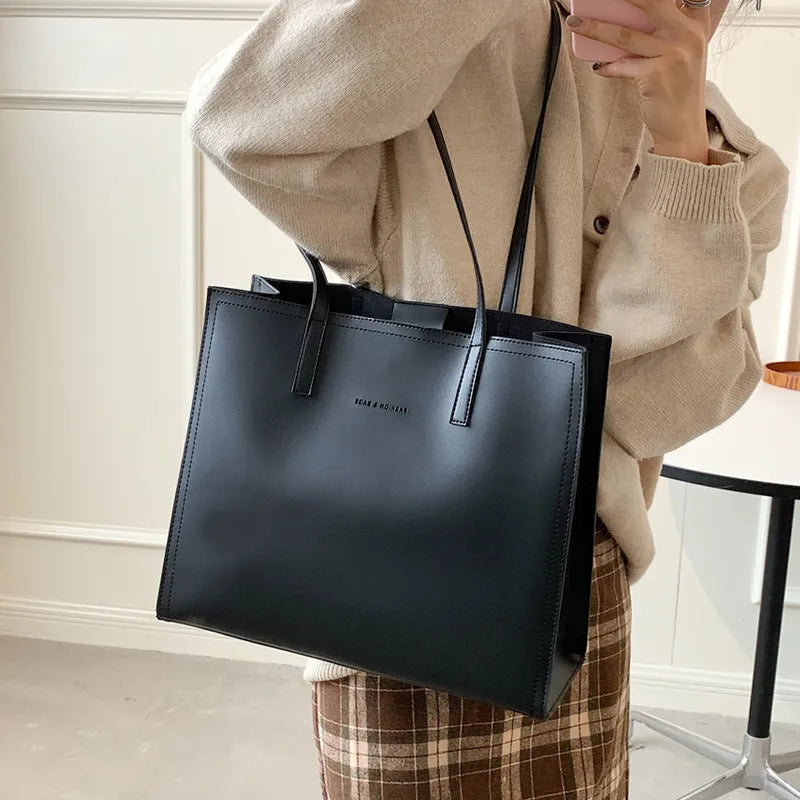 Sac à main noir en cuir porté par une femme en pull beige et jupe à carreaux. Accessoire mode élégant, tendance automne, style chic et minimaliste.