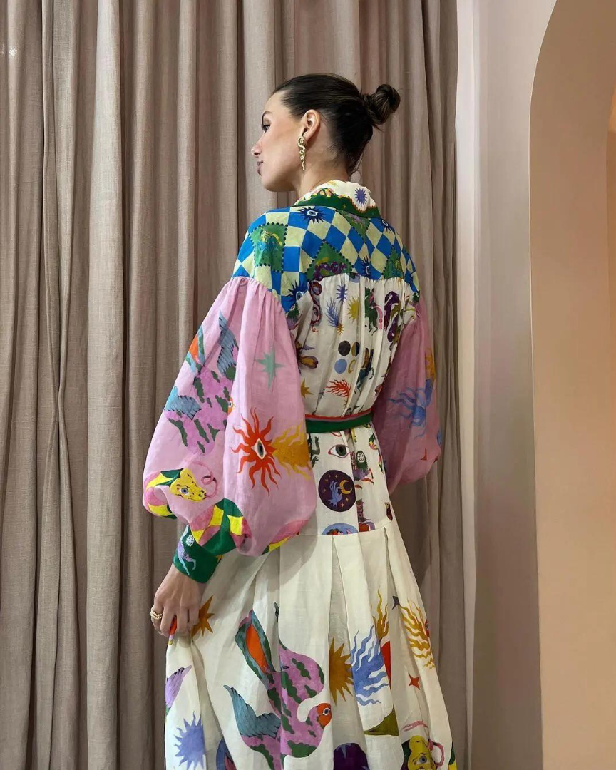 Femme portant une robe colorée avec motifs géométriques et artistiques, manches bouffantes roses, devant un rideau beige. Mode tendance et originale.