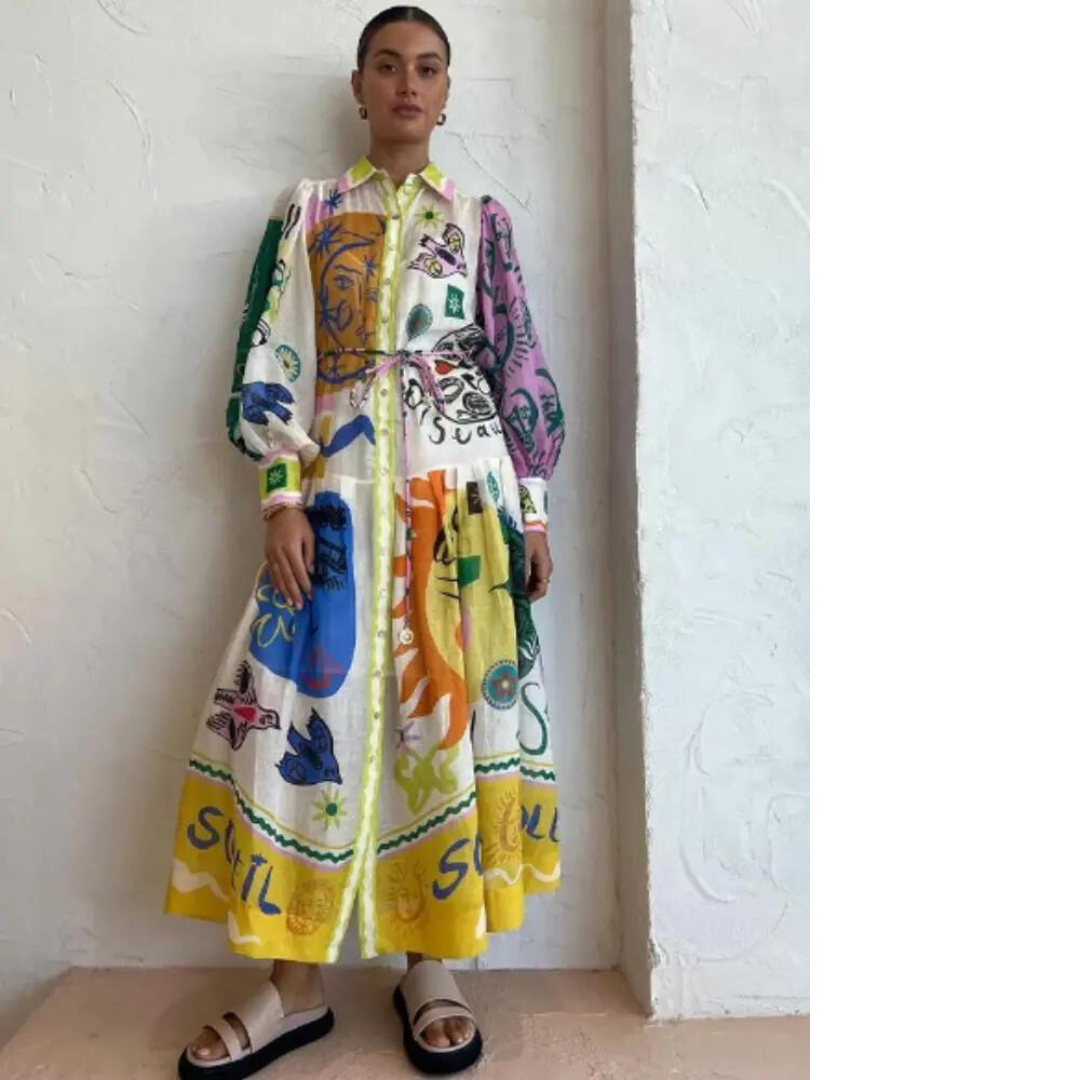 Robe longue colorée avec motifs artistiques, manches longues, portée par une femme debout contre un mur blanc. Mode féminine, style bohème chic.