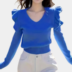 Femme portant un pull bleu à manches longues avec volants, col en V, associé à un pantalon blanc. Mode féminine élégante et tendance.