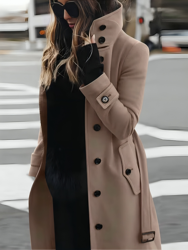 Femme élégante en manteau beige à boutons, lunettes de soleil noires, traversant la rue. Mode automne chic, style urbain sophistiqué.