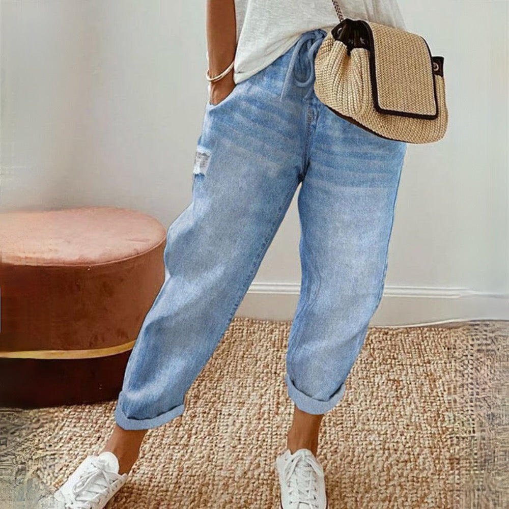 Femme portant un jean décontracté bleu clair, baskets blanches et sac à main en paille, debout sur un tapis beige. Mode décontractée chic.