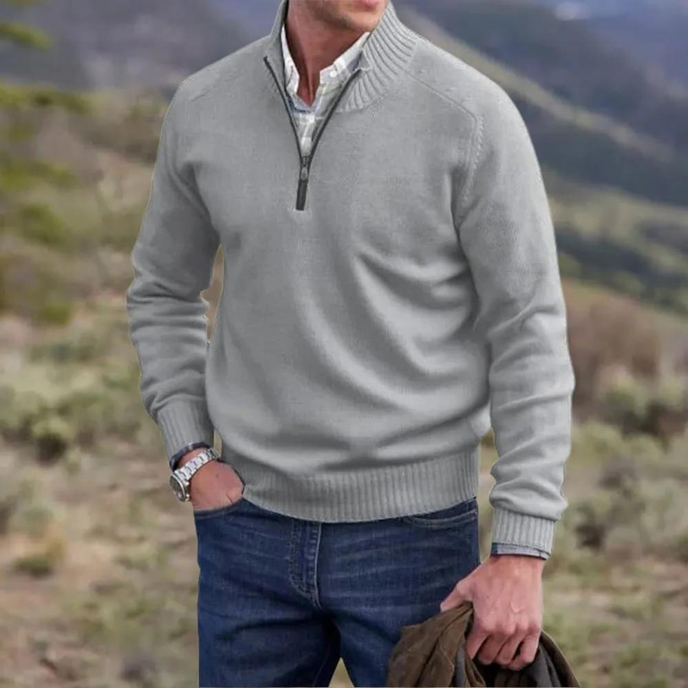 Homme portant un pull gris à col zippé, chemise blanche et jean bleu, debout en extérieur avec paysage flou en arrière-plan. Mode masculine décontractée.