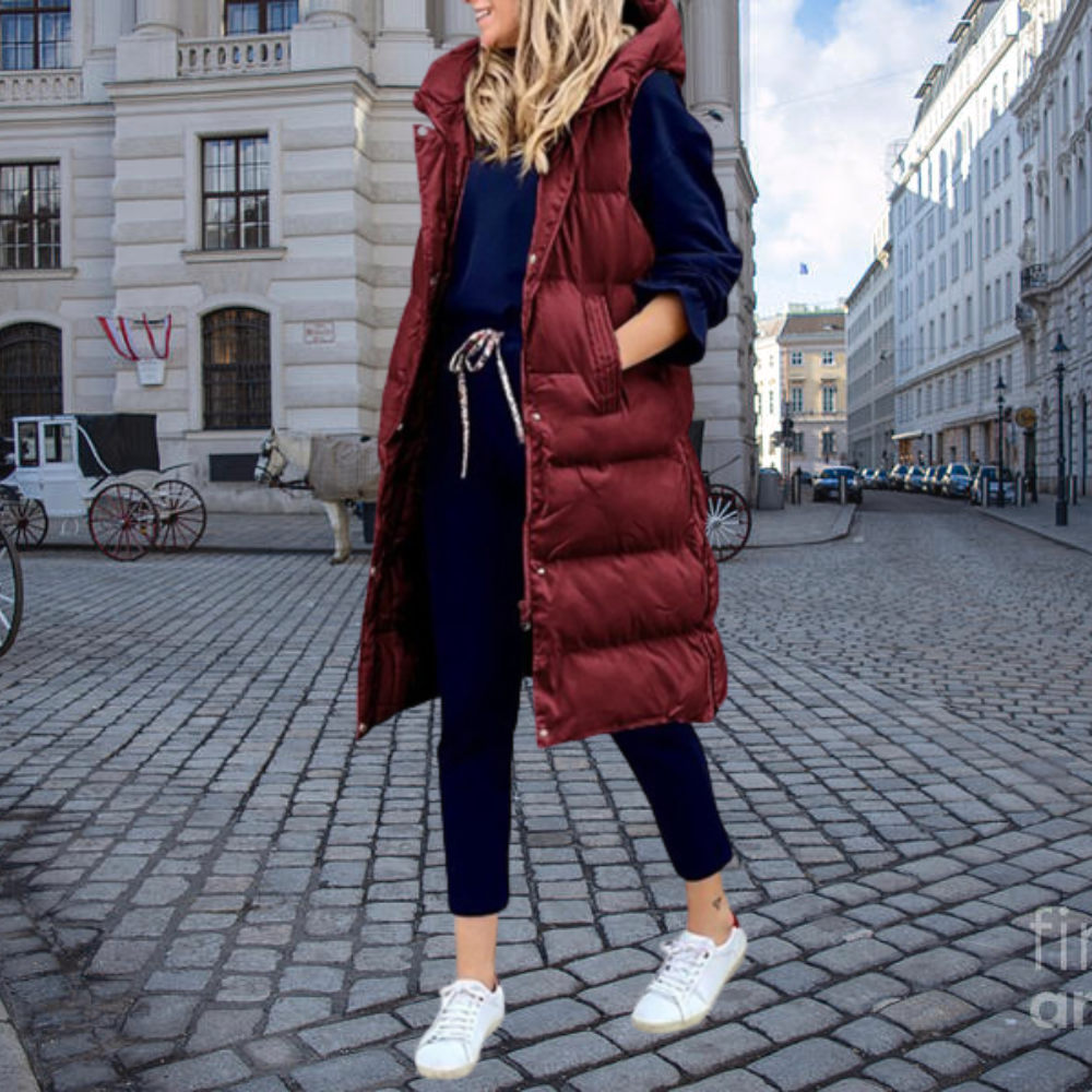 Femme en manteau long bordeaux, tenue décontractée, rue pavée, architecture européenne, journée ensoleillée, mode urbaine, sneakers blanches.